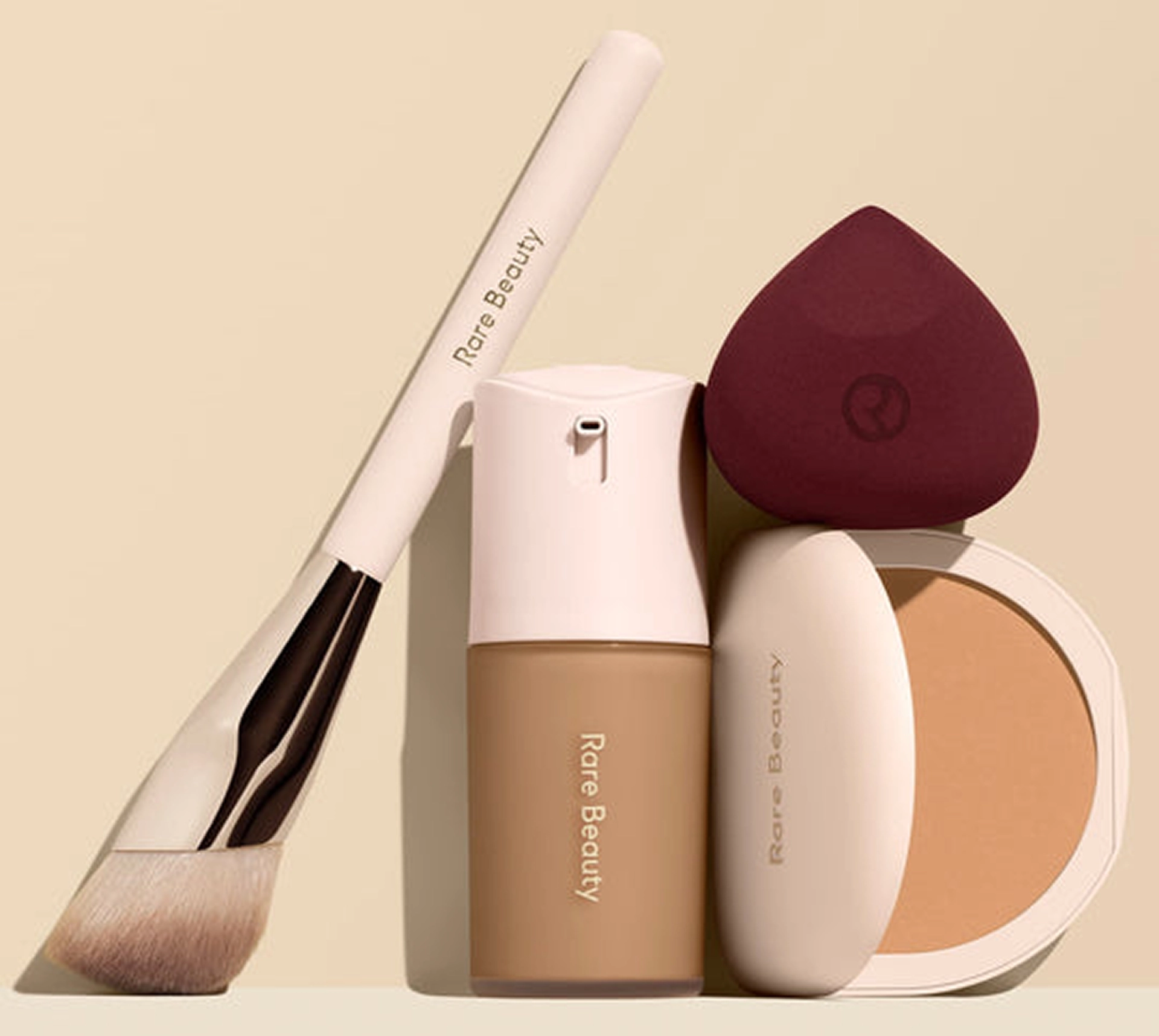 Rare Beauty complexion products arranged against a cream background: an angled face brush, a pump-top liquid foundation in a warm tan shade, a burgundy teardrop makeup sponge, and an open pressed powder compact.