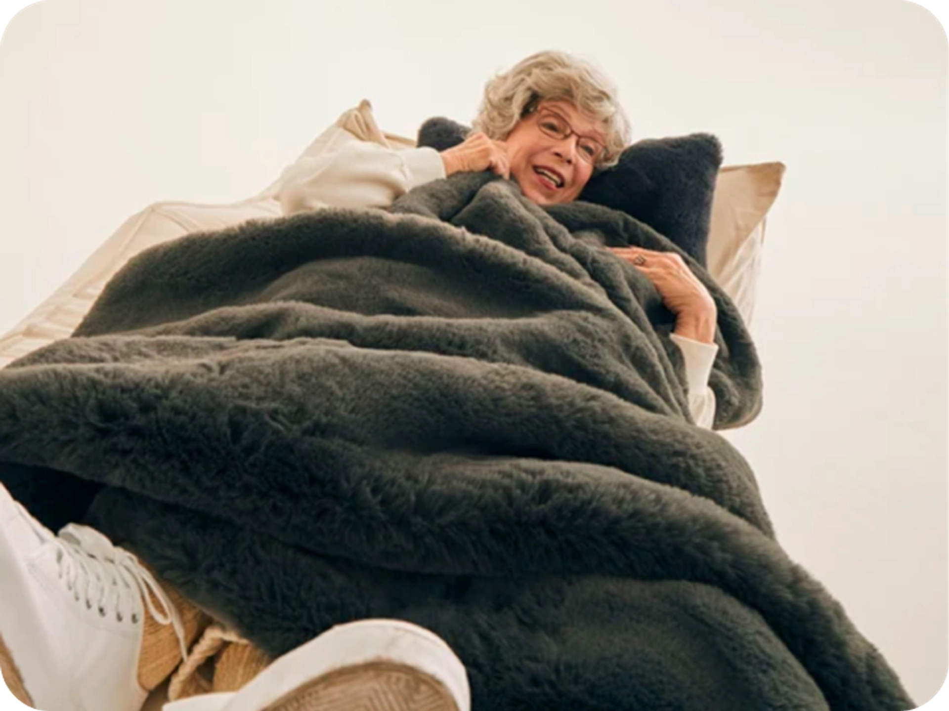 Low-angle view of a smiling older woman with short gray hair and glasses reclining under a charcoal faux fur blanket, with her white sneakers in the foreground.
