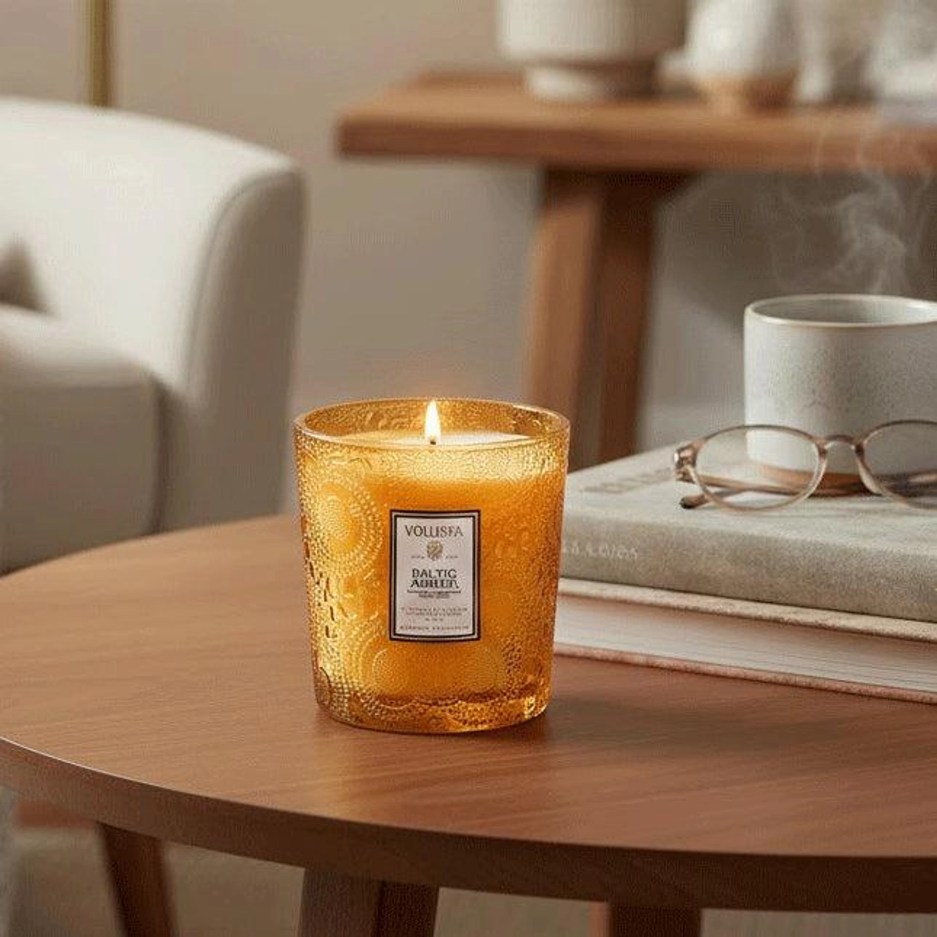Voluspa Baltic Amber candle in an embossed amber glass jar, lit and sitting on a round wooden coffee table beside a stack of books topped with eyeglasses and a steaming mug.