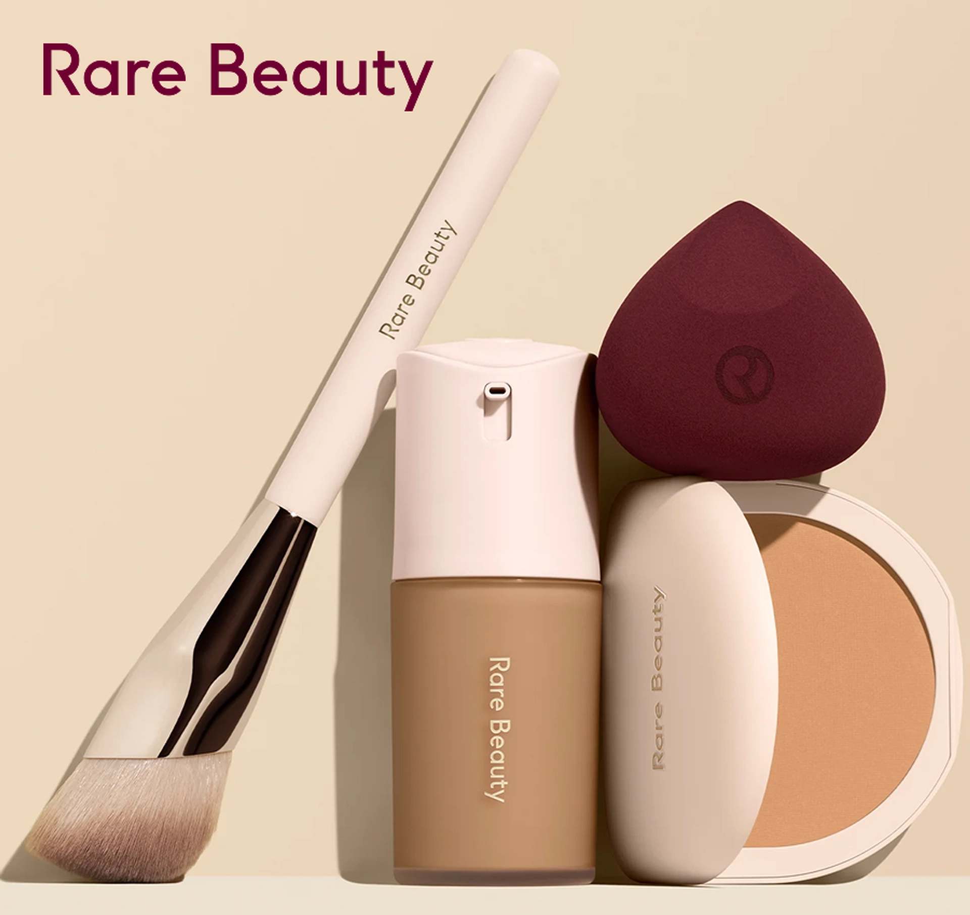 Rare Beauty complexion products arranged against a cream background: an angled face brush, a pump-top liquid foundation in a warm tan shade, a burgundy teardrop makeup sponge, and an open pressed powder compact.