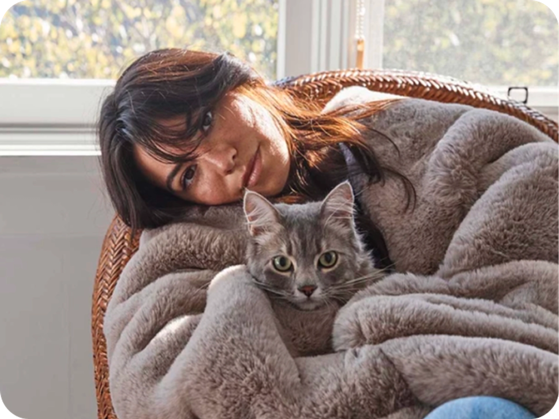 Smiling woman resting her head on her arm in a woven rattan chair, wrapped in a taupe faux fur blanket with a gray tabby cat on her lap, in front of a sunlit window.