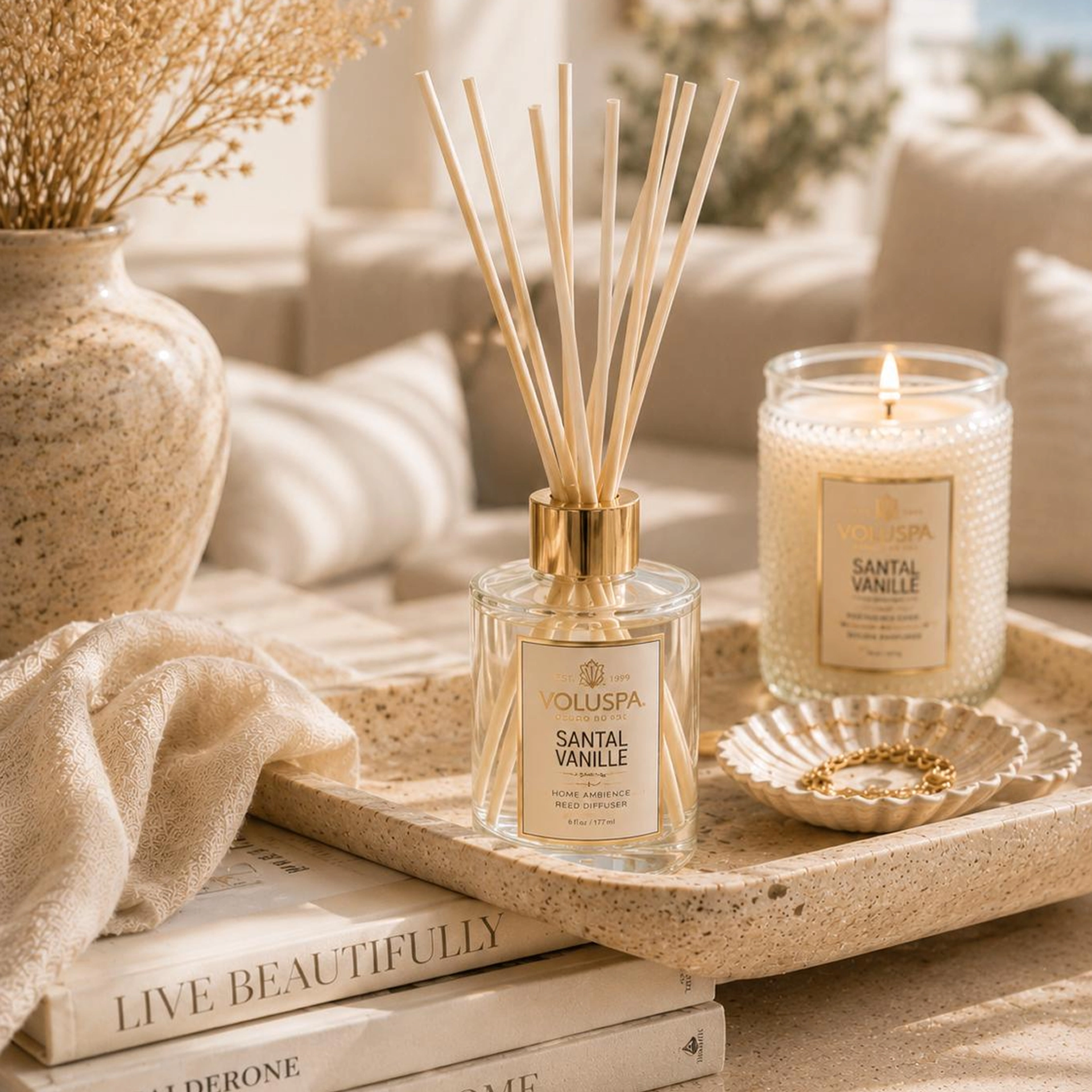 Voluspa Santal Vanille home ambience reed diffuser with a gold collar and natural reeds, styled on a speckled stone tray beside a lit Santal Vanille candle, a scalloped dish holding a gold chain bracelet, and a stack of books in a cream-toned living room.