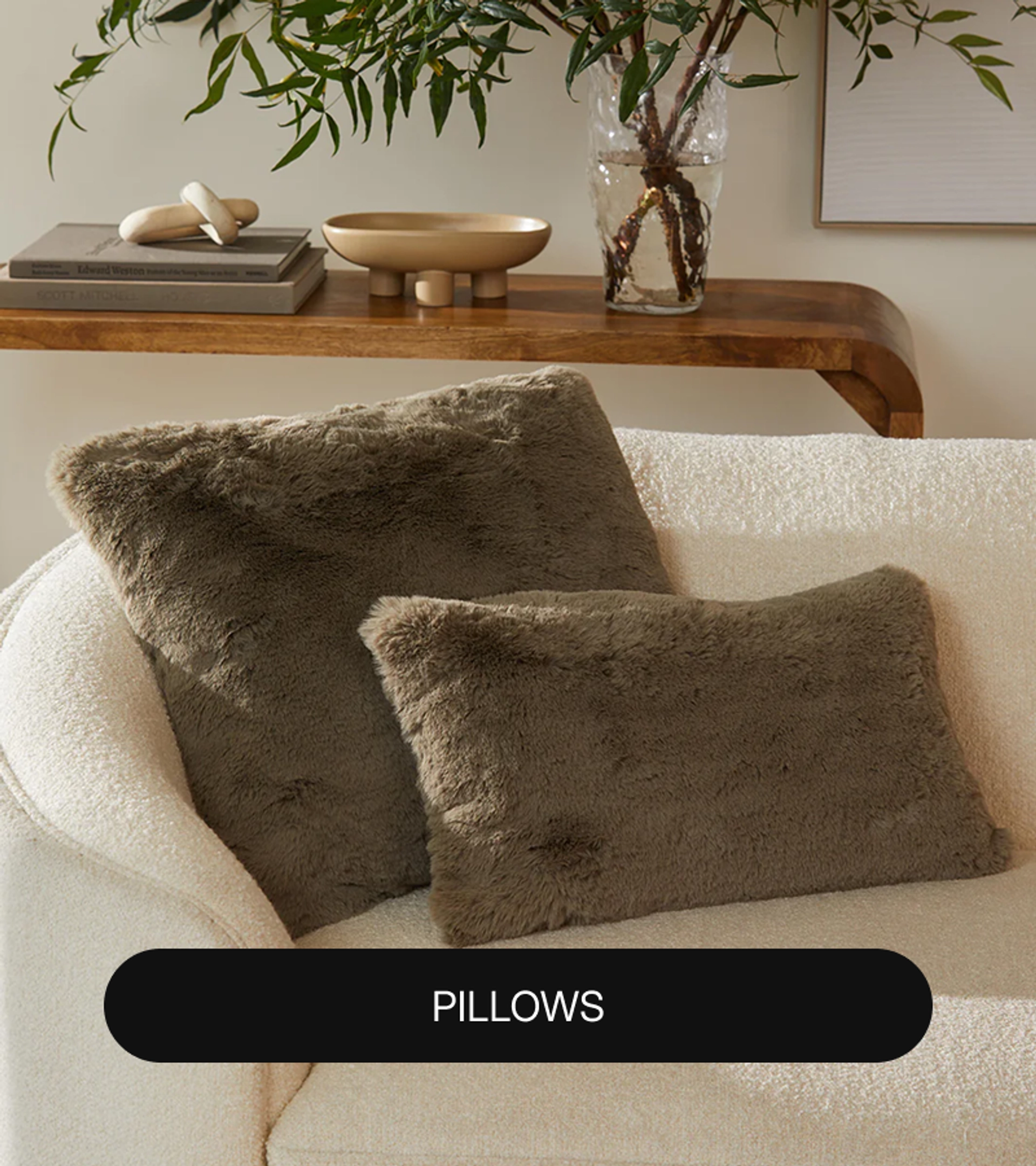 Two olive-green faux fur pillows, one square and one rectangular, on a cream bouclé sofa in front of a wooden console table with books, a bowl, and a vase of greenery, with a "Pillows" label at the bottom.