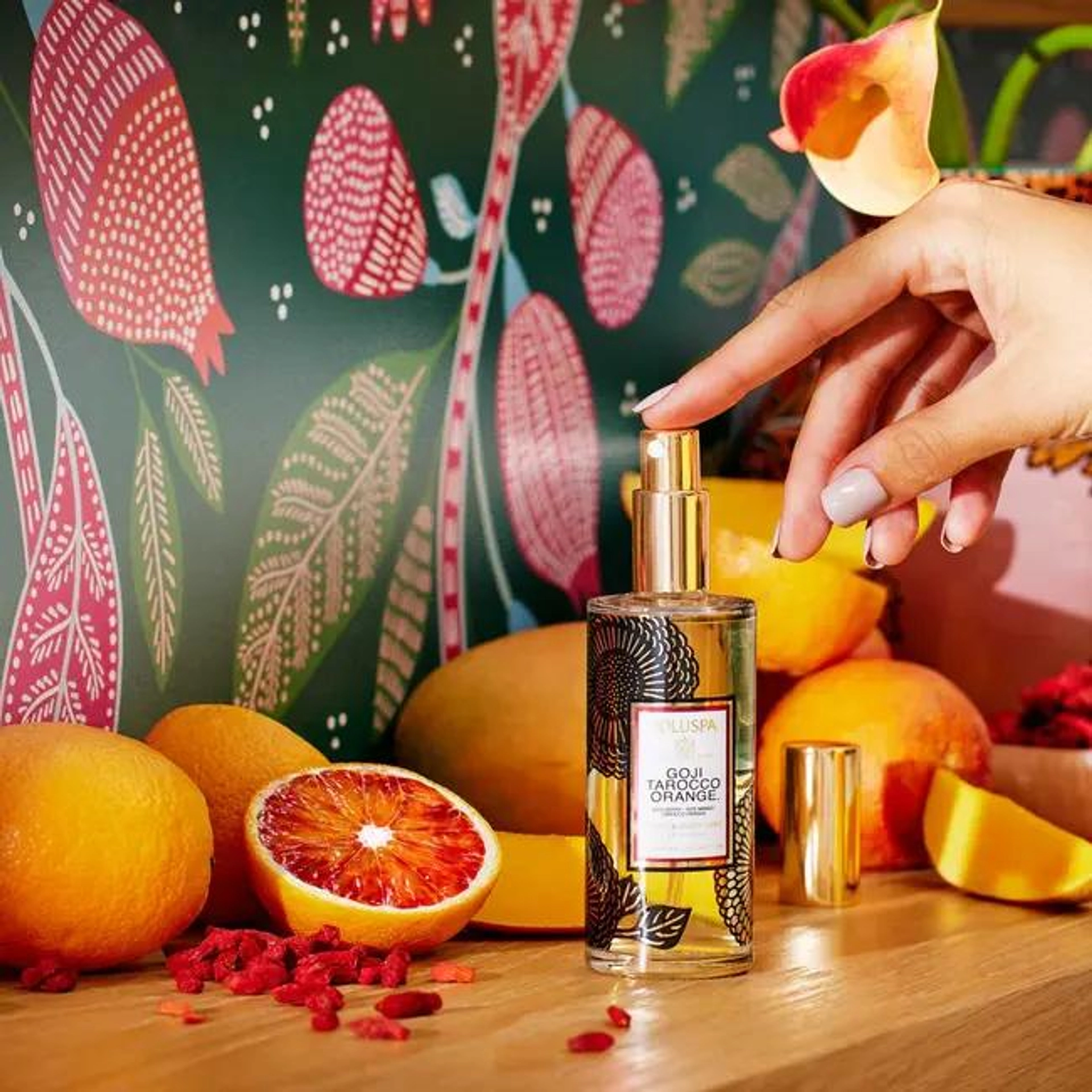 A hand pressing the gold pump of a Voluspa Goji Tarocco Orange room and body mist in a glass bottle with a black floral label, surrounded by blood oranges, mangoes, and scattered goji berries on a wooden shelf in front of a colorful botanical mural.