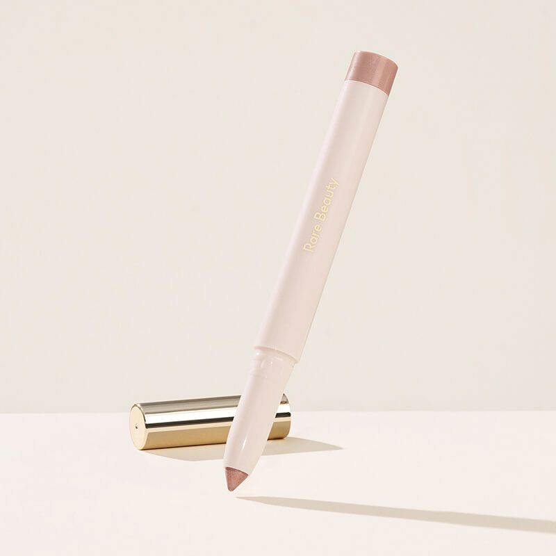 Rare Beauty All of the Above Weightless Eyeshadow Stick in shade Well-Being, a cream-colored twist-up pen with a shimmery rose-taupe tip, shown upright and angled beside its gold cap on a cream background.
