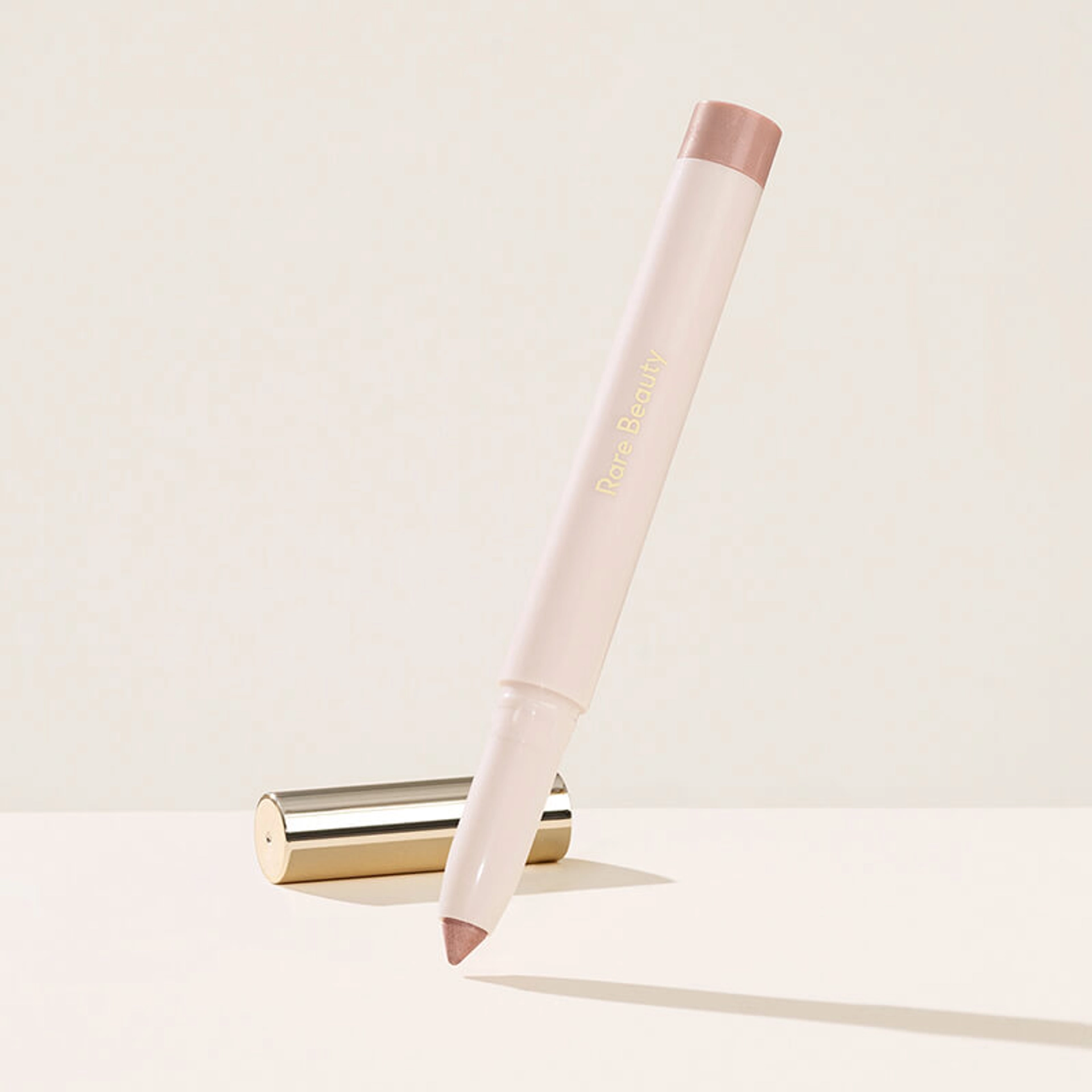 Rare Beauty All of the Above Weightless Eyeshadow Stick in shade Well-Being, a cream-colored twist-up pen with a shimmery rose-taupe tip, shown upright and angled beside its gold cap on a cream background.