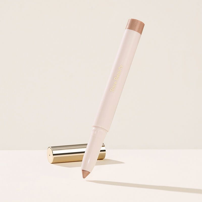 Rare Beauty All of the Above Weightless Eyeshadow Stick in shade Integrity, a cream-colored twist-up pen with a shimmery champagne-pink tip, shown upright and angled beside its gold cap on a cream background.