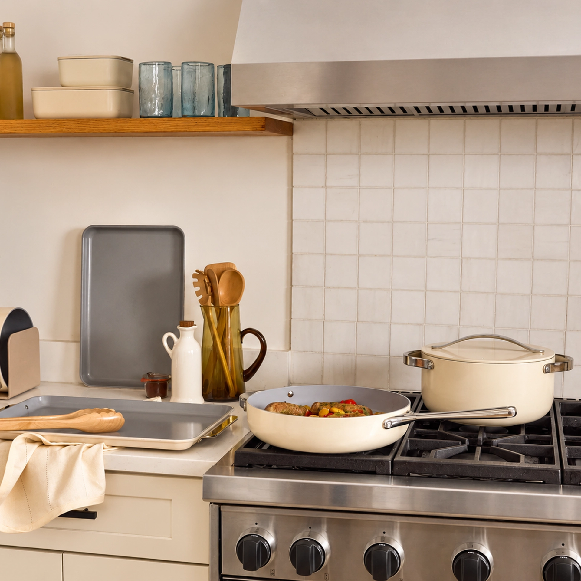 Caraway cream cookware in a kitchen, with a frying pan of sausages and peppers and a lidded pot on a stainless steel gas range, beside a gray baking sheet, wooden utensils in a pitcher, and cream storage containers on a shelf.