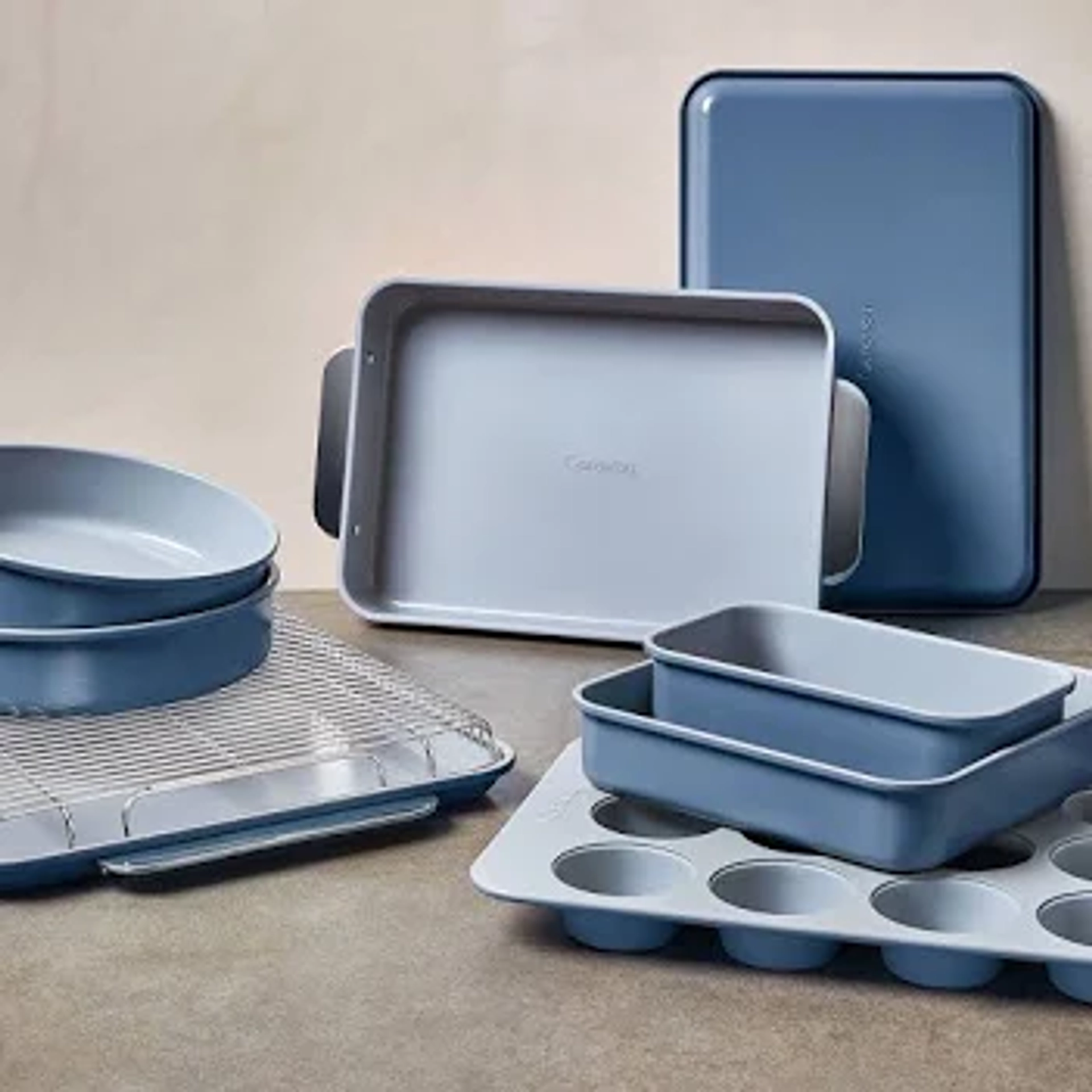 Caraway ceramic-coated bakeware set in slate blue, including a rectangular baking pan, round cake pans, a loaf pan, a muffin tin, a cooling rack, and a large baking sheet, arranged on a gray surface.