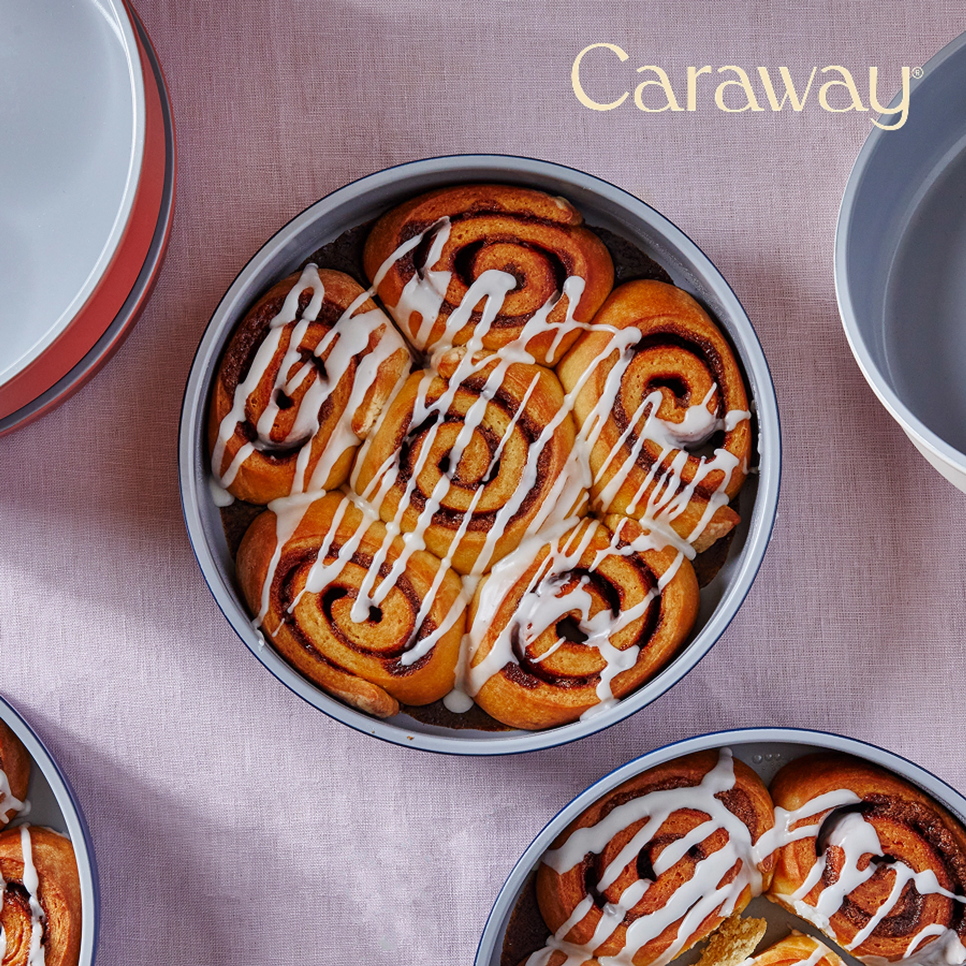 Overhead view of glazed cinnamon rolls baked in a gray ceramic-coated round Caraway cake pan with a navy rim, surrounded by more pans of rolls and stacked empty pans in gray and terracotta on a lavender linen background.