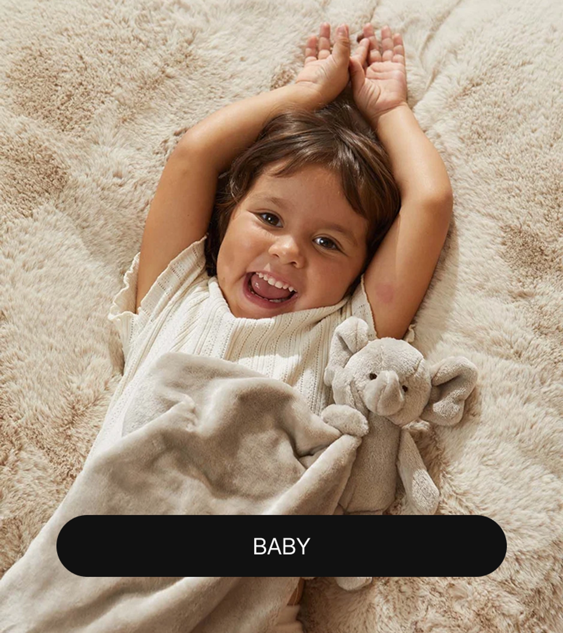 Laughing toddler lying on a cream faux fur surface with arms raised overhead, holding a gray plush elephant lovey blanket, with a "Baby" label at the bottom.