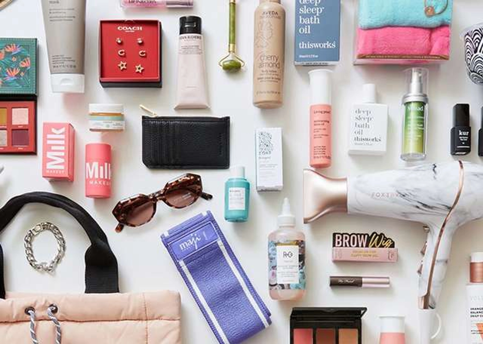 Flat lay of beauty, skincare, and lifestyle products on a white surface, including a marble hair dryer, a cherry almond lotion, bath oil boxes, Milk Makeup items, a gold stud earring set in a red Coach box, tortoiseshell sunglasses, a black cardholder, a jade facial roller, a purple resistance band, and a quilted peach tote bag.