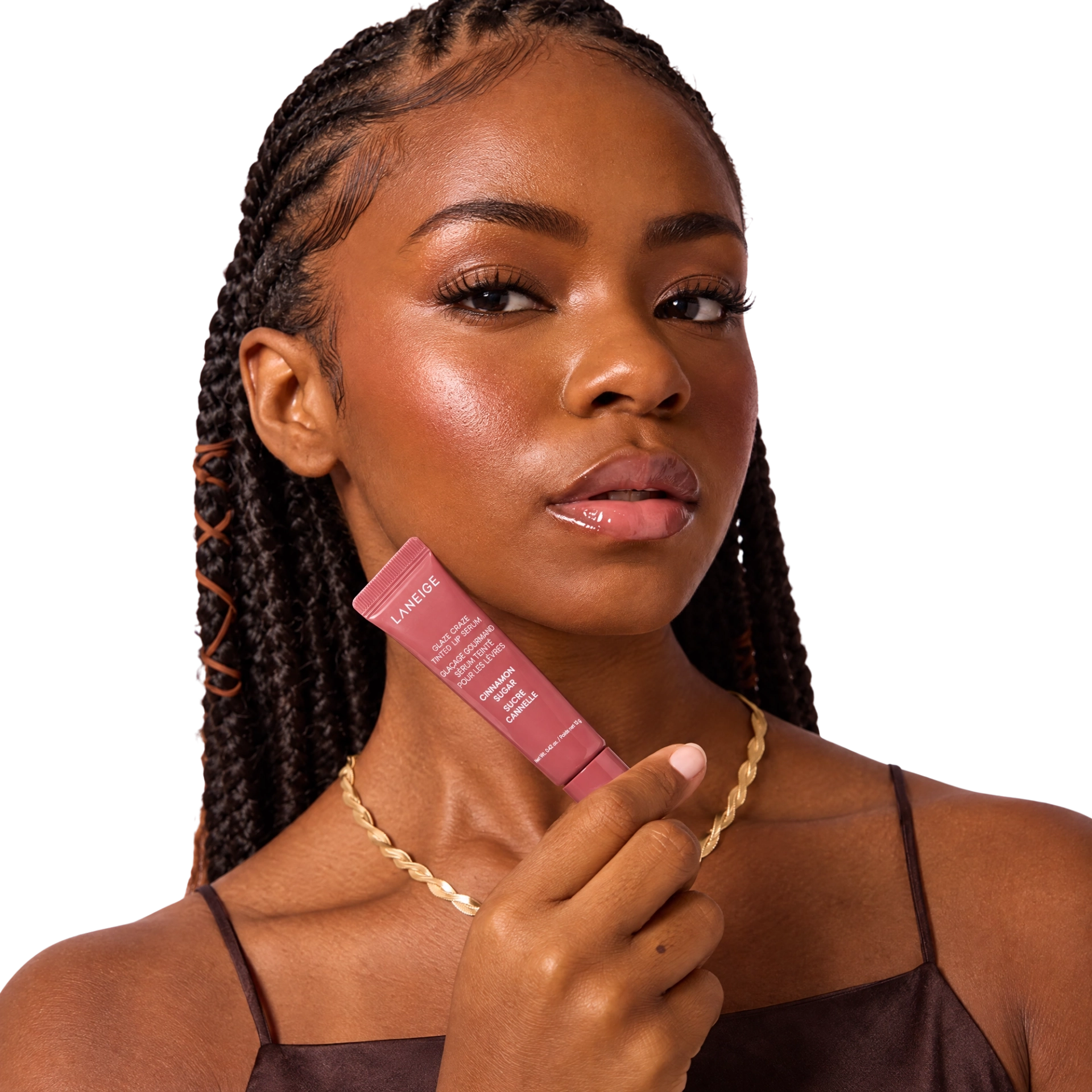 A Black woman with braided hair holds a Laneige Glaze Craze Tinted Lip Serum in Cinnamon Sugar near her cheek, showcasing glossy, radiant skin and a natural makeup look.