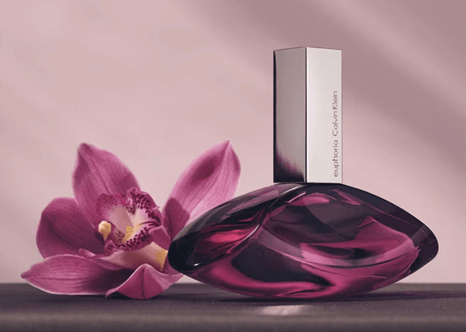 Calvin Klein Euphoria perfume in a curved, deep magenta glass bottle with a silver rectangular cap, resting beside a pink orchid on a dark surface against a soft mauve background.