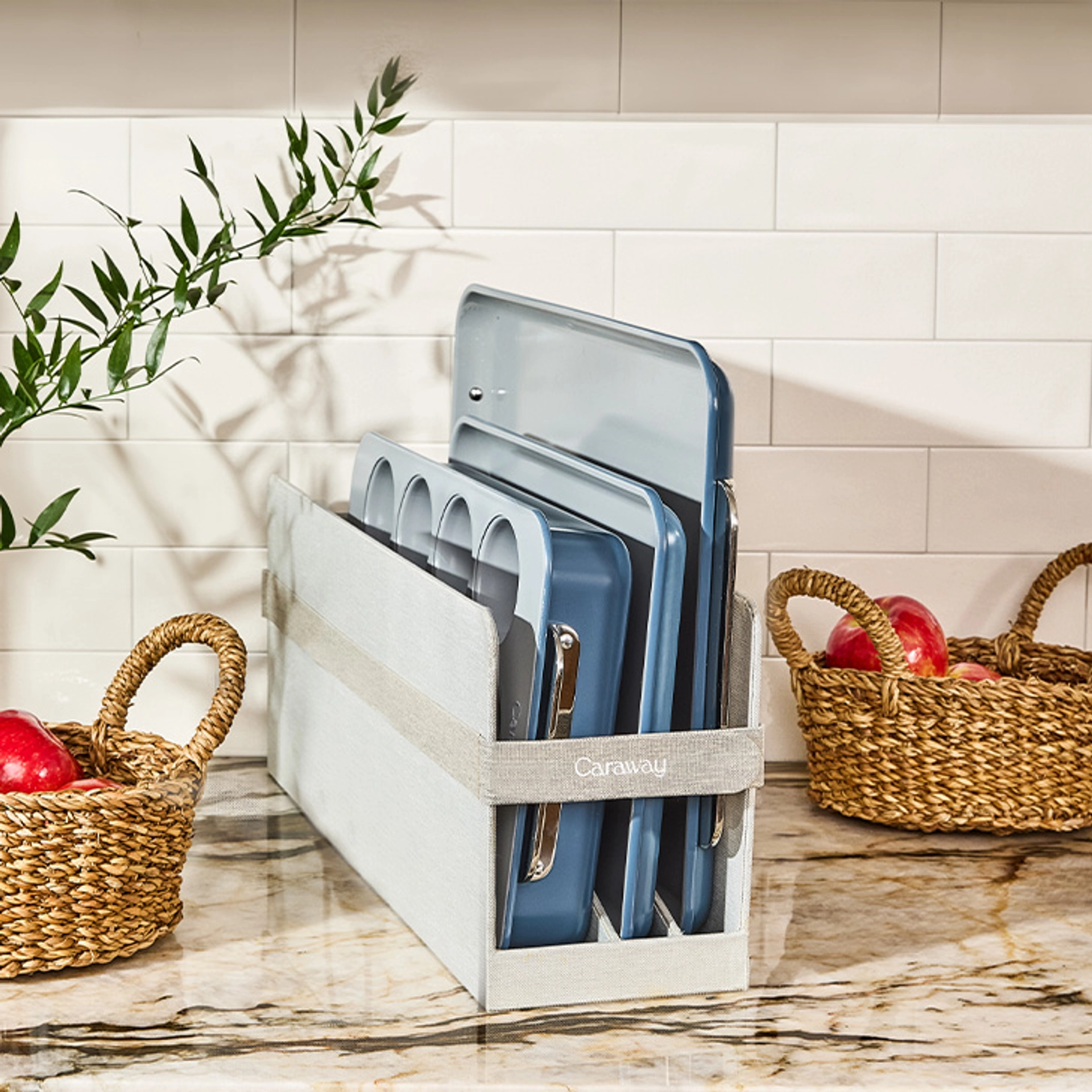 Caraway slate blue bakeware, including a muffin tin and baking sheets with stainless steel handles, stored upright in a gray fabric Caraway organizer on a marble kitchen counter beside two woven baskets of red apples and a leafy green branch.