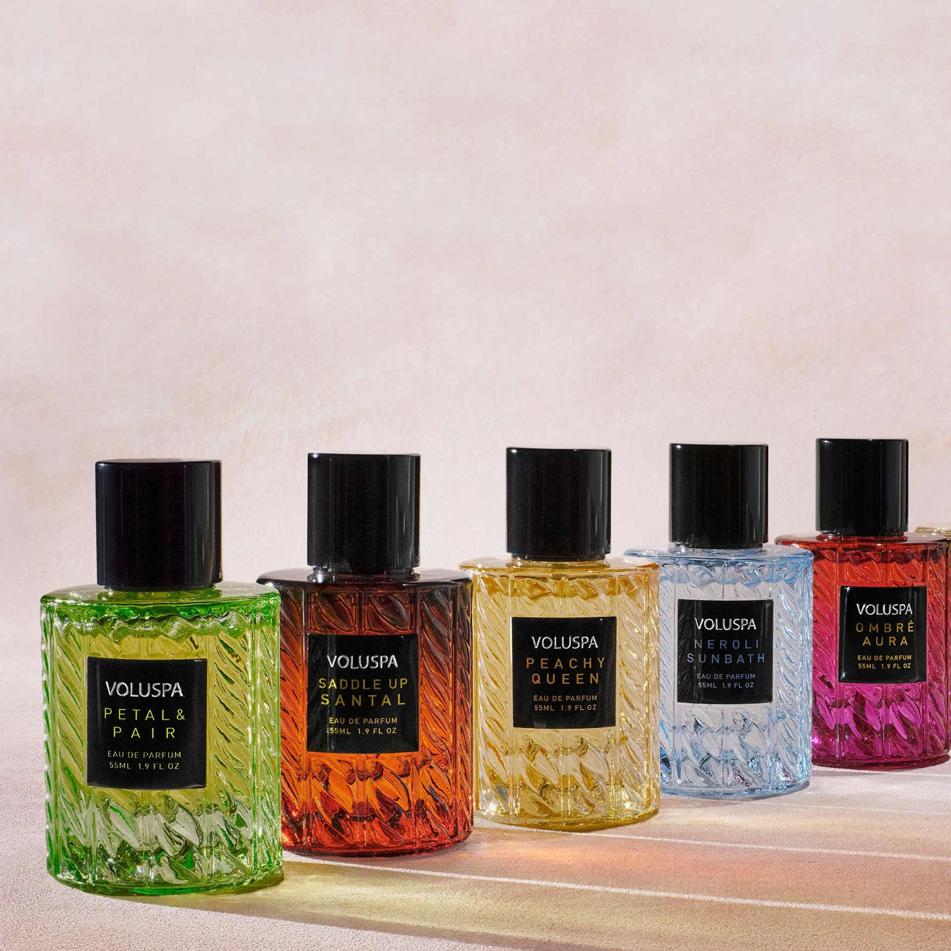 Row of five Voluspa Eau de Parfum bottles with black caps and textured glass in green, amber, yellow, light blue, and magenta-red, labeled Petal & Pair, Saddle Up Santal, Peachy Queen, Neroli Sunbath, and Ombré Aura, lined up against a soft pink background.