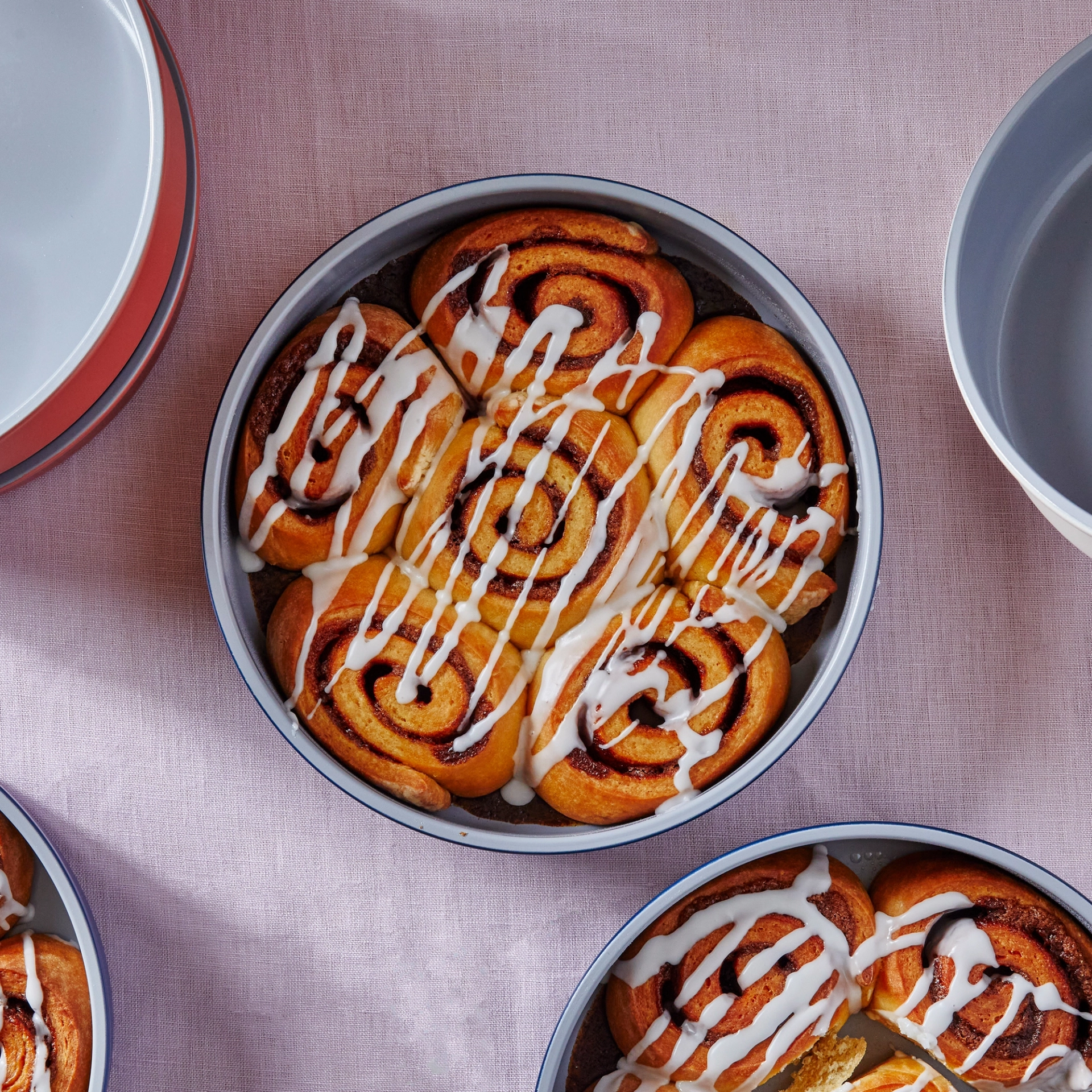Overhead view of glazed cinnamon rolls baked in a gray ceramic-coated round Caraway cake pan with a navy rim, surrounded by more pans of rolls and stacked empty pans in gray and terracotta on a lavender linen background.