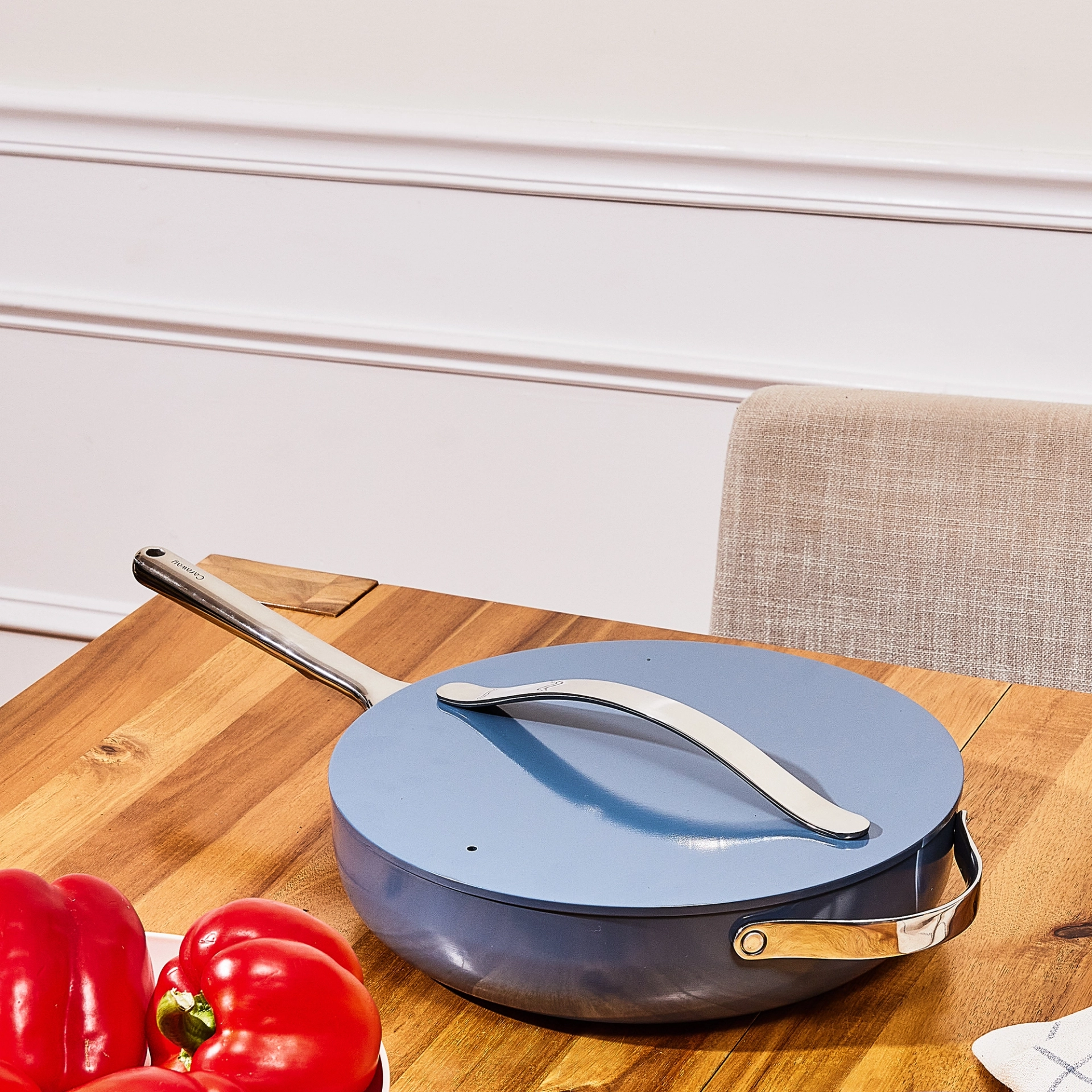 Caraway slate blue sauté pan with its lid on and a stainless steel handle, sitting on a wooden table beside a bowl of red bell peppers, with a linen-upholstered chair and white paneled wall in the background.