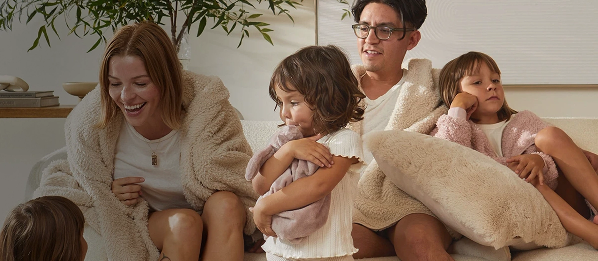 A family relaxing together on a cream bouclé sofa in cozy UnHide faux fur: a laughing woman and a man in glasses wrapped in cream sherpa robes, a toddler hugging a mauve plush lovey, and a child in a pink fuzzy jacket leaning on a large cream faux fur pillow.