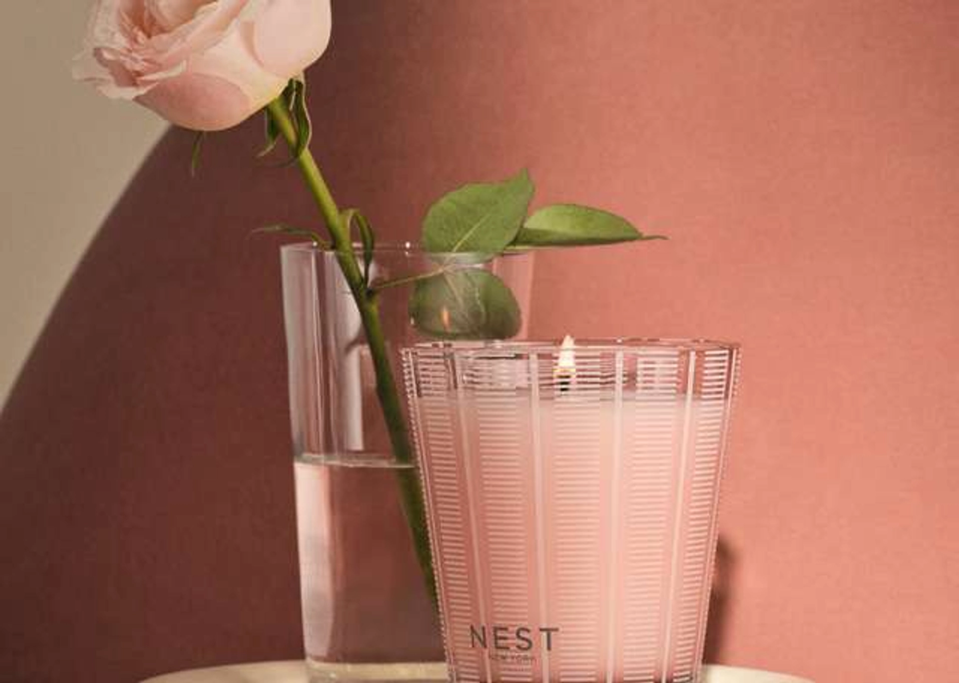 Nest New York frosted glass candle with a vertical stripe pattern, lit and placed in front of a glass vase holding a single pale pink rose, against a dusty rose wall.