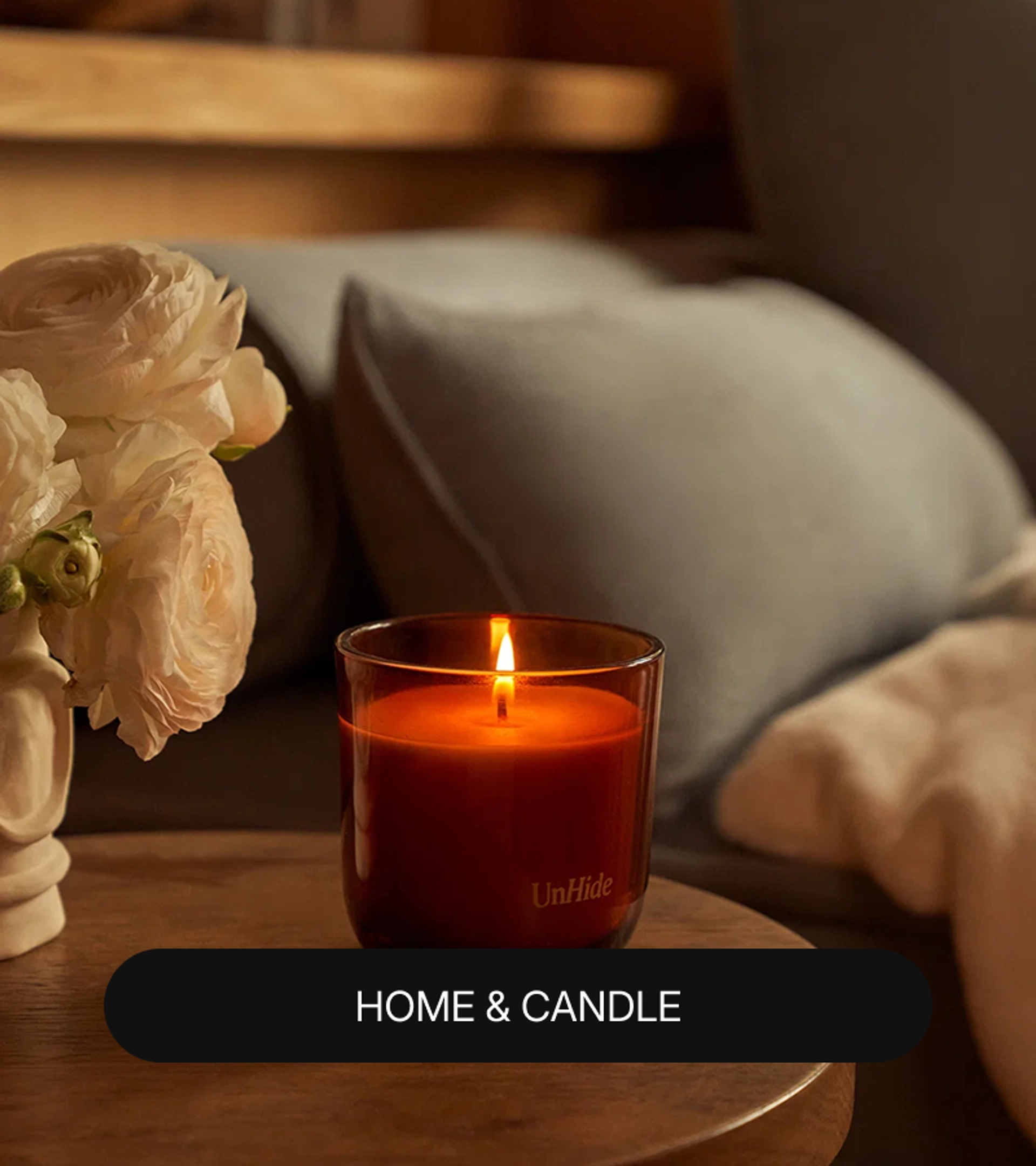 Lit amber glass UnHide candle on a round wooden side table beside a vase of white ranunculus flowers, with a gray pillow and cream throw in the background and a "Home & Candle" label at the bottom.