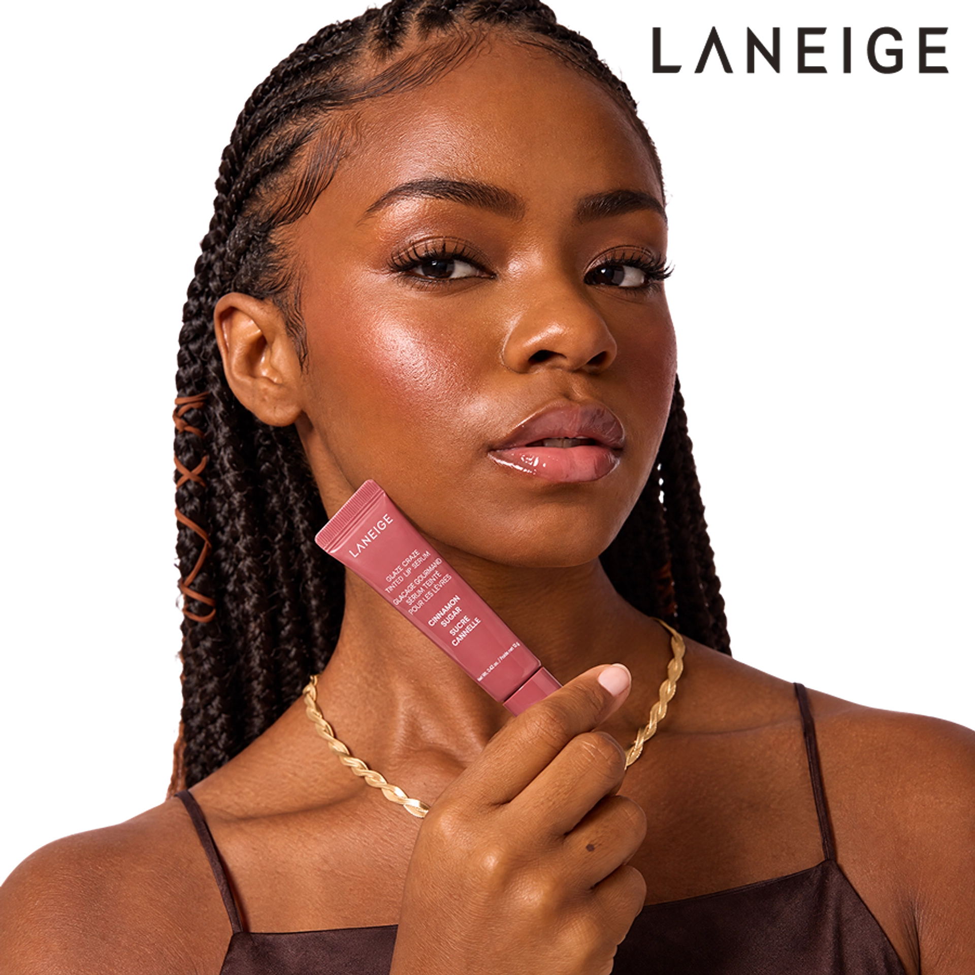 A Black woman with braided hair holds a Laneige Glaze Craze Tinted Lip Serum in Cinnamon Sugar near her cheek, showcasing glossy, radiant skin and a natural makeup look.
