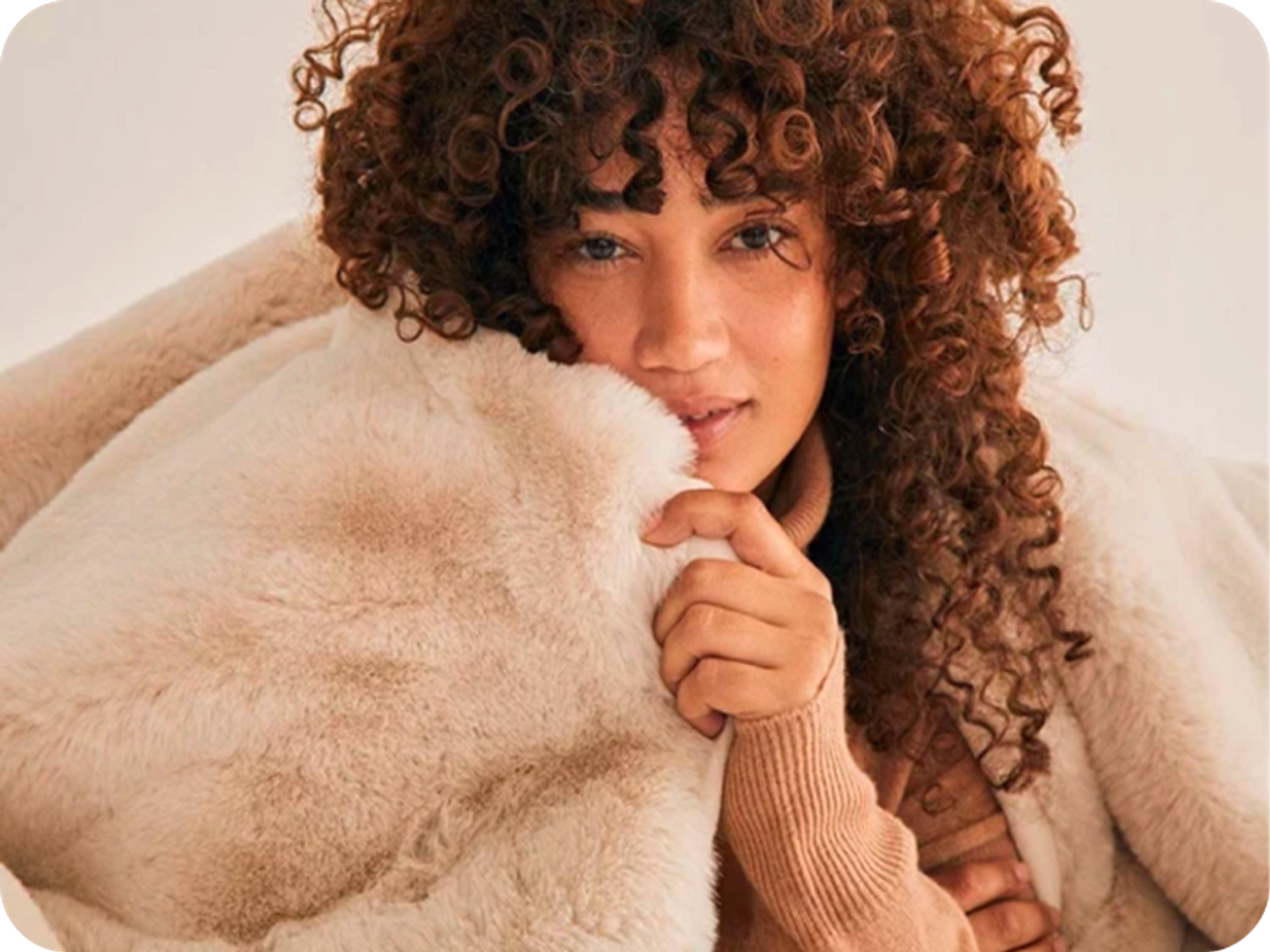 Woman with curly auburn hair and a tan ribbed sweater snuggled into a cream faux fur blanket, holding the edge up to her cheek.